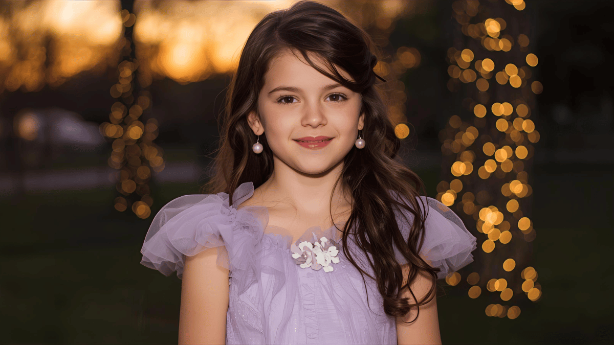 A girl in a lavender tulle dress with floral details, loose curls, pearl earrings, and soft pink makeup, smiling at sunset near twinkling fairy lights.