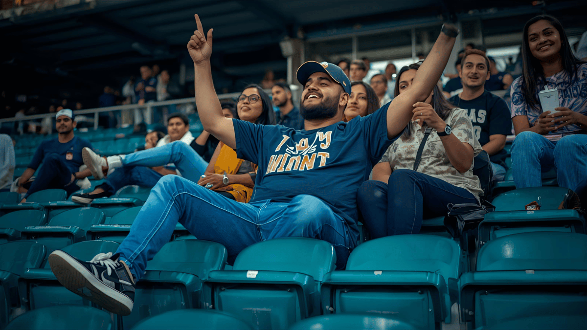 A man wearing jeans, a team t-shirt, sneakers, and a baseball cap, cheering in the stands with friends.