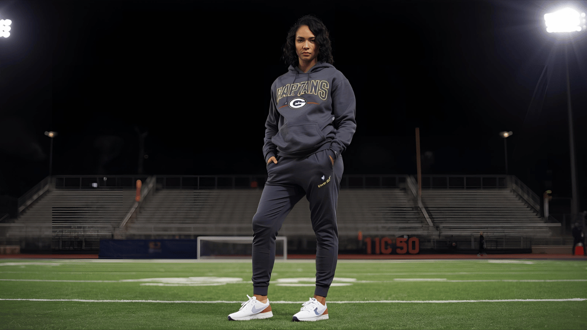 A woman at a night game wearing joggers, sneakers, and a team sweatshirt, with stadium lights behind her.