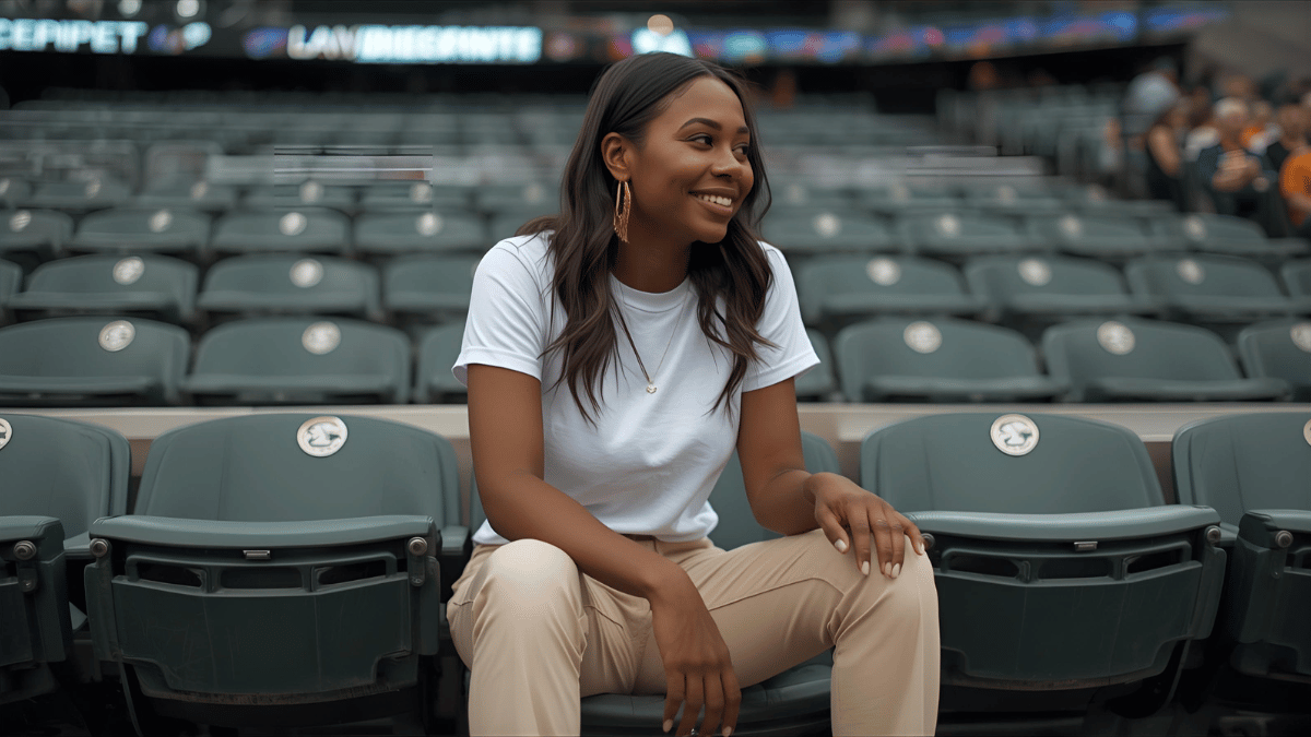A woman sitting in a baseball field seat, wearing beige trousers, a white tee, and gold hoops, smiling while watching the game.