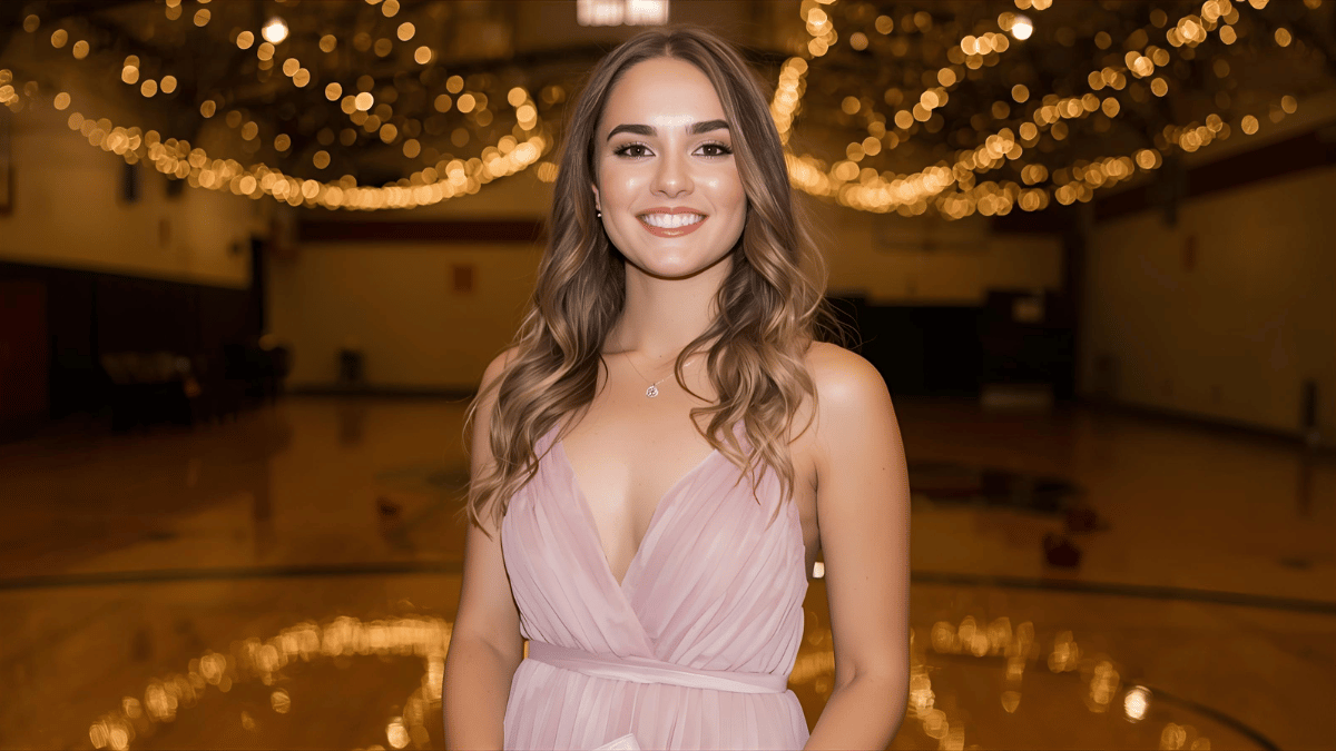 A young woman standing under fairy lights in a high school gym, wearing a blush pink chiffon homecoming dress, soft curls, natural makeup, and holding a small clutch while smiling confidently.