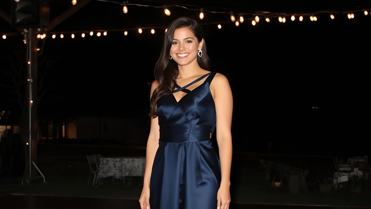 A young woman wearing a navy fit-and-flare satin dress, silver heels, and elegant earrings, standing under soft string lights at a homecoming dance.