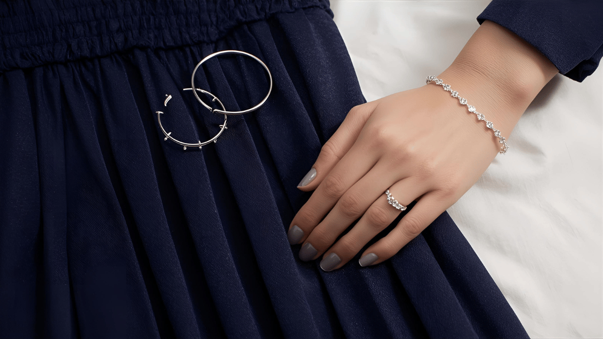 Close-up of delicate silver jewelry — hoops, bracelet, and a dainty ring — placed beside a navy-blue dress on a soft fabric background.